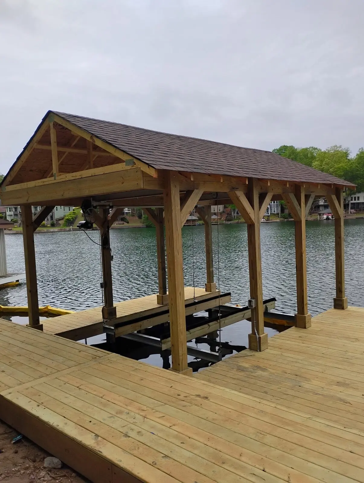 Timber-framed boathouse with a shingled gable roof over a boat lift and open slip, dock decking still unstained and no rail installed yet