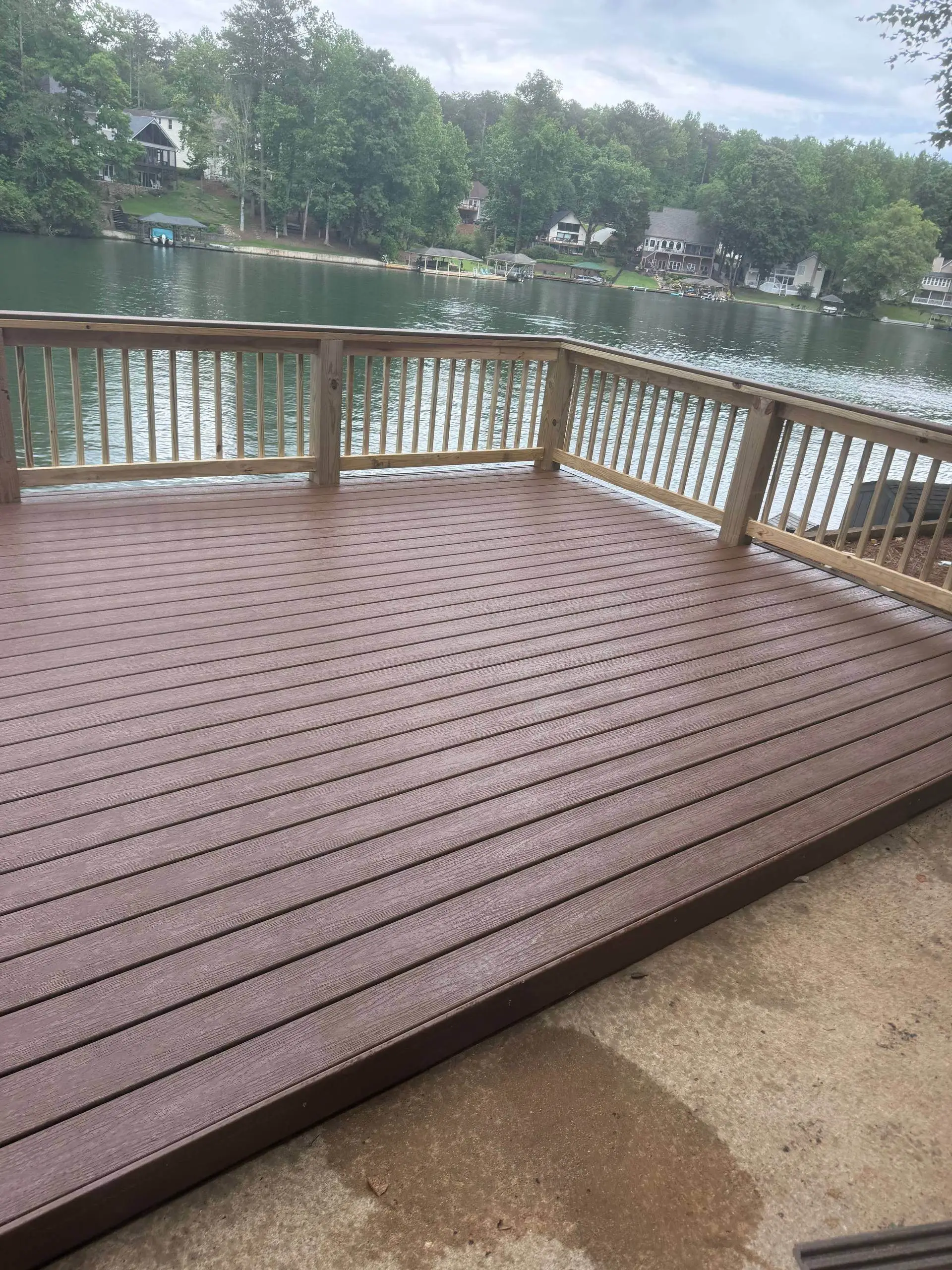 Finished composite deck with a natural-tone (unstained) wood rail, lake and tree-lined shoreline view, workers visible on the ground below near the water's edge.