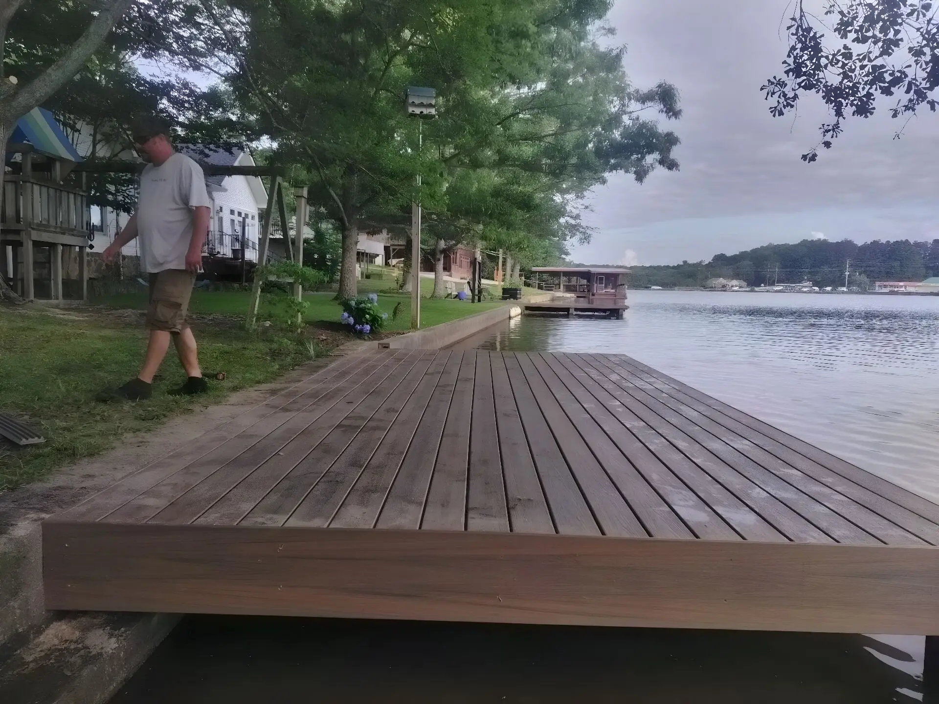 New composite dock platform built at the water's edge, a man walking the bank beside it, a covered dock further down the same shore and lake homes behind