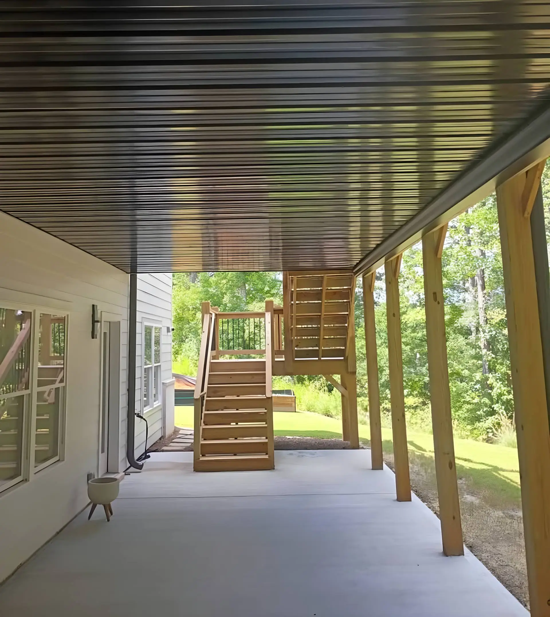 Covered patio with a black corrugated metal ceiling and concrete floor, wood staircase leading up to an elevated deck with a black metal rail, backyard lawn, pool, and trees visible beyond
