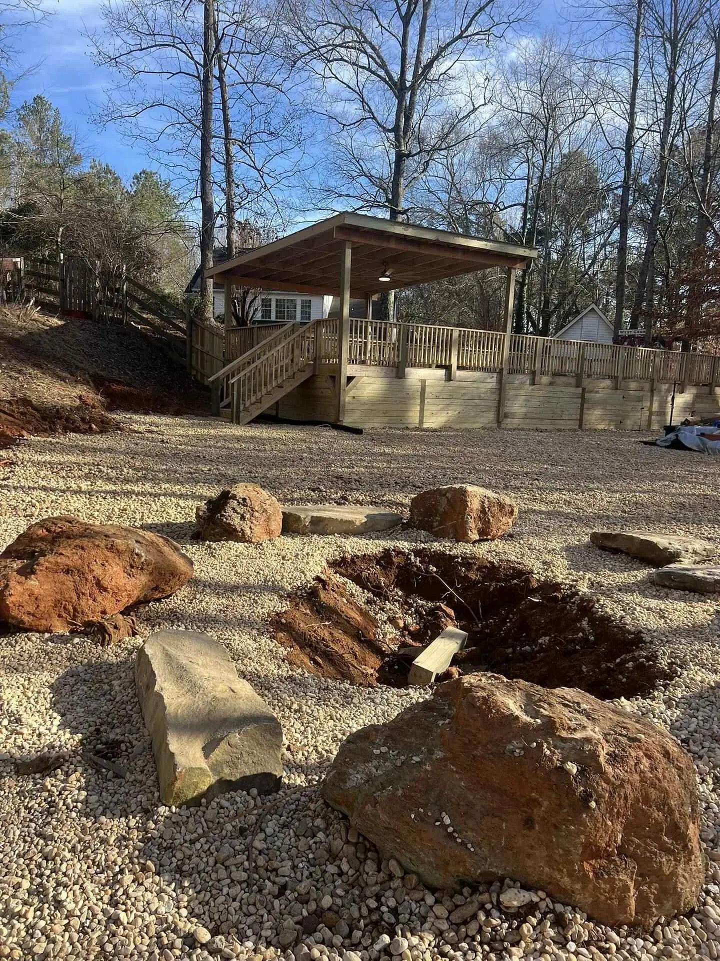 Elevated deck with an open-sided roof and ceiling fan, wood stairs and a timber retaining wall, boulders and an open hole in the gravel in front
