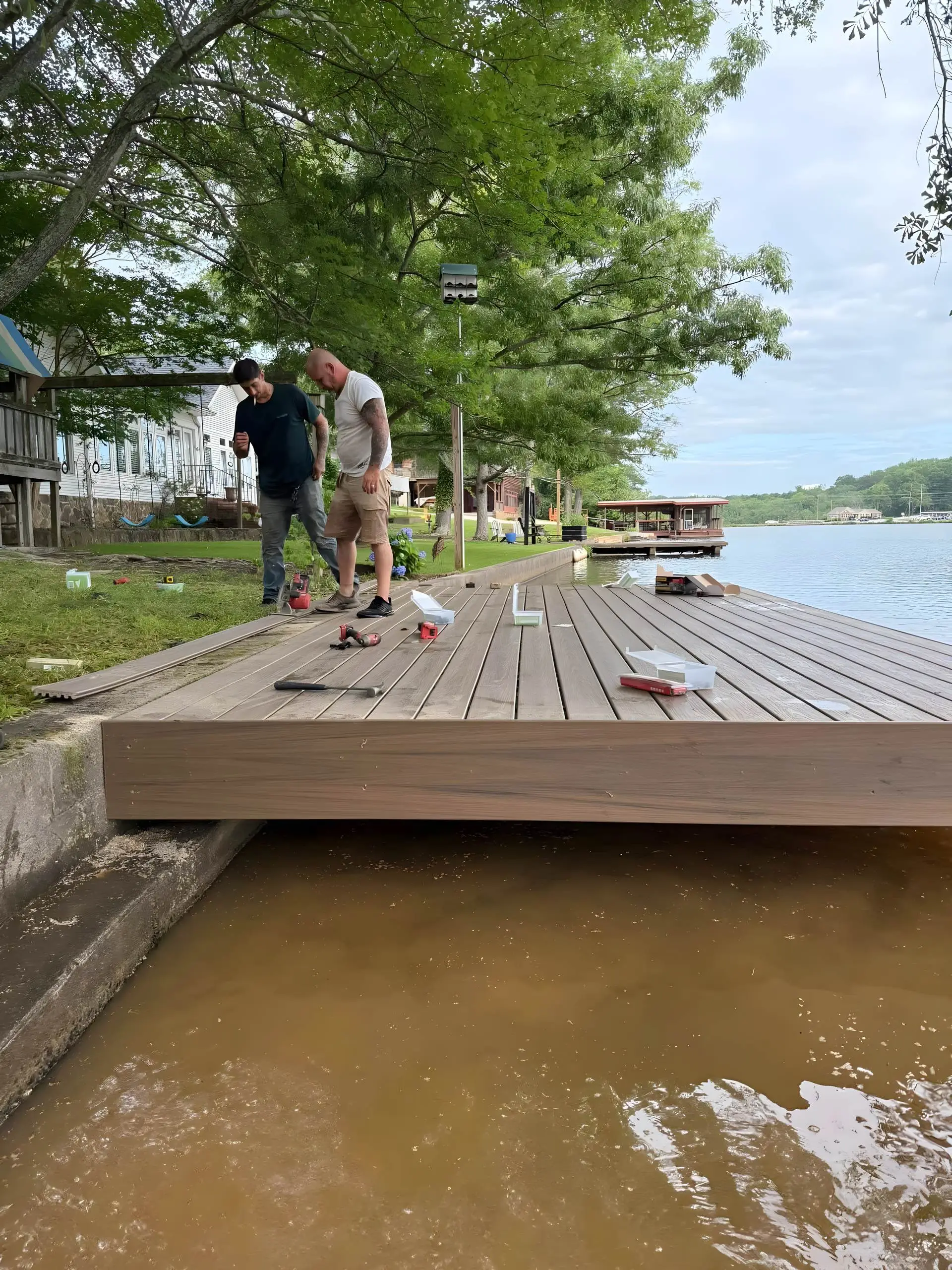Two crew members installing composite decking at the edge of a seawall, tools and hardware boxes scattered on the deck, water below.