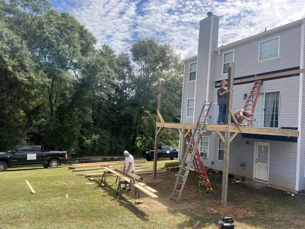 Crew framing an elevated second-story deck on a two-story home, ladders and lumber staged in the yard