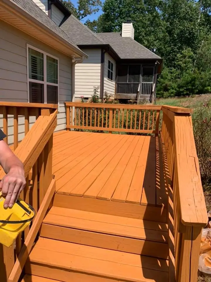 Close detail of a hand applying deck stain from a yellow pail onto fresh-toned deck boards