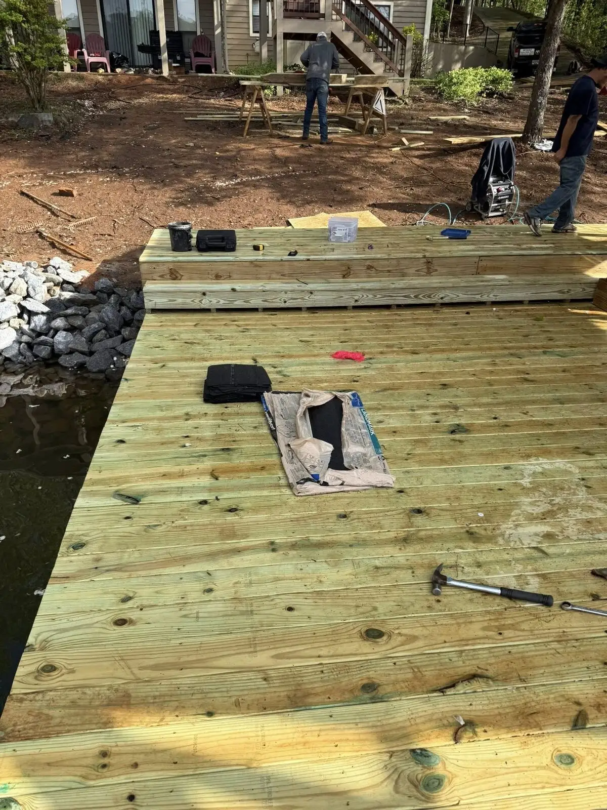 New pressure-treated dock decking freshly laid along a rock riprap seawall, crew cutting boards on sawhorses on shore