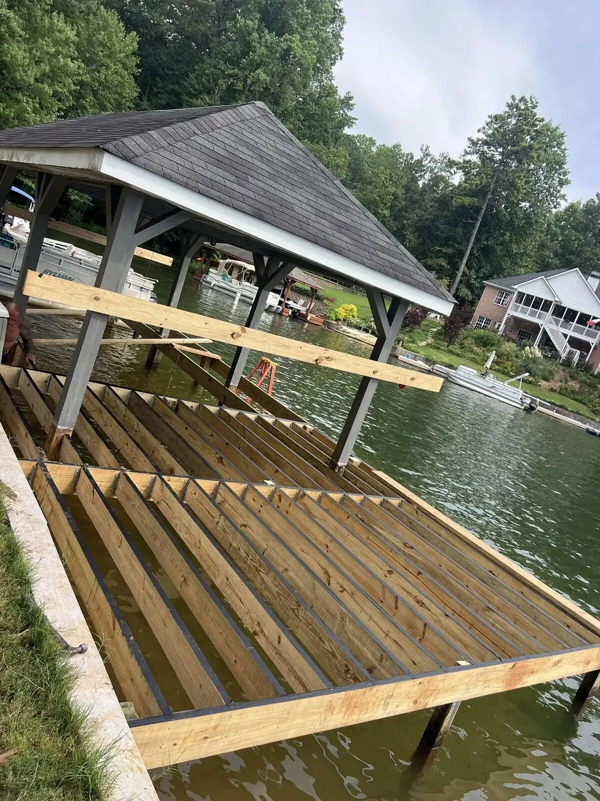 Bare pressure-treated joists of a new dock walkway extending over lake water, no decking laid yet, covered boathouse alongside