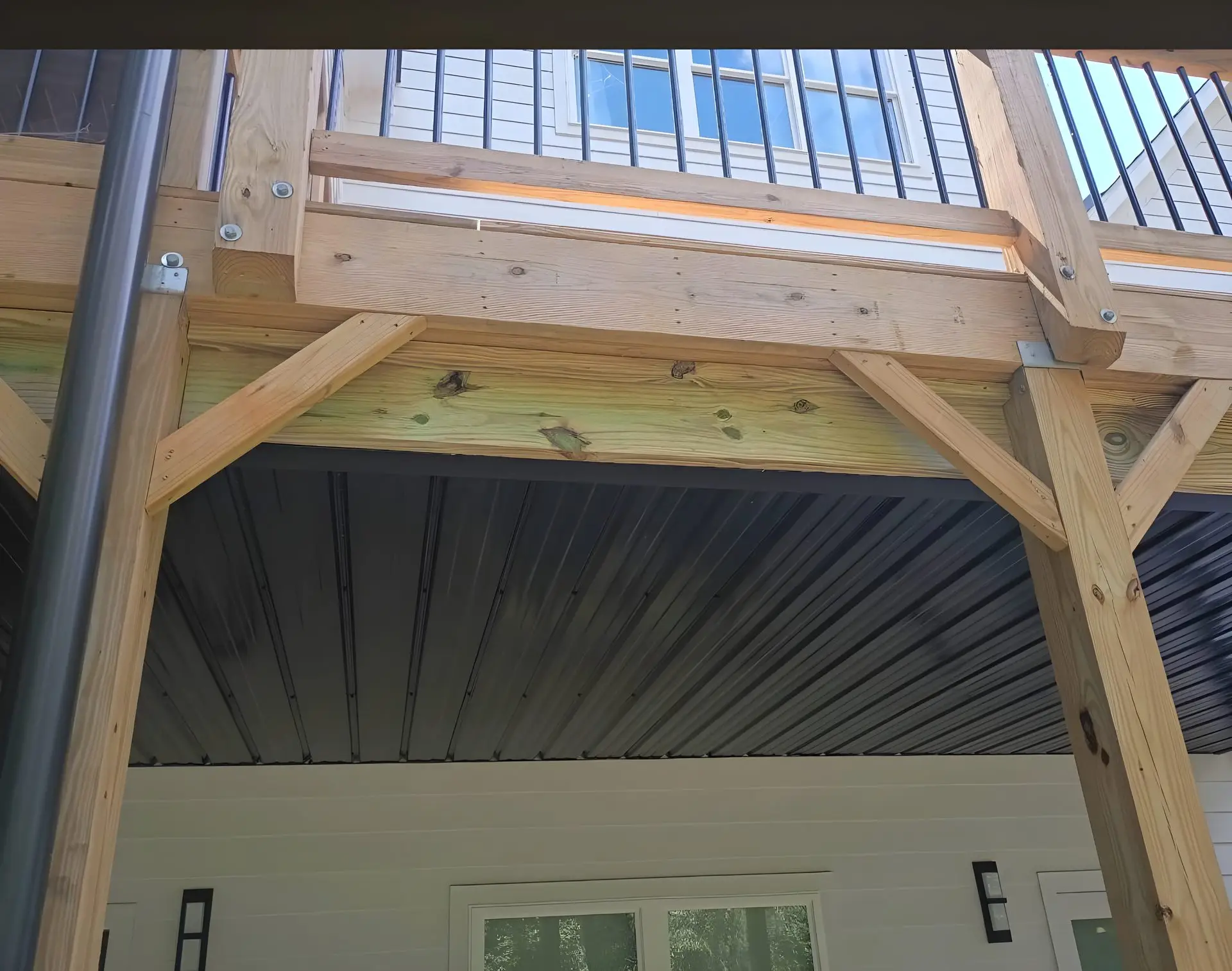 Underside structural framing of an elevated deck: pressure-treated posts and beams supporting the deck above, black corrugated metal ceiling panel underneath, house siding and a window visible.