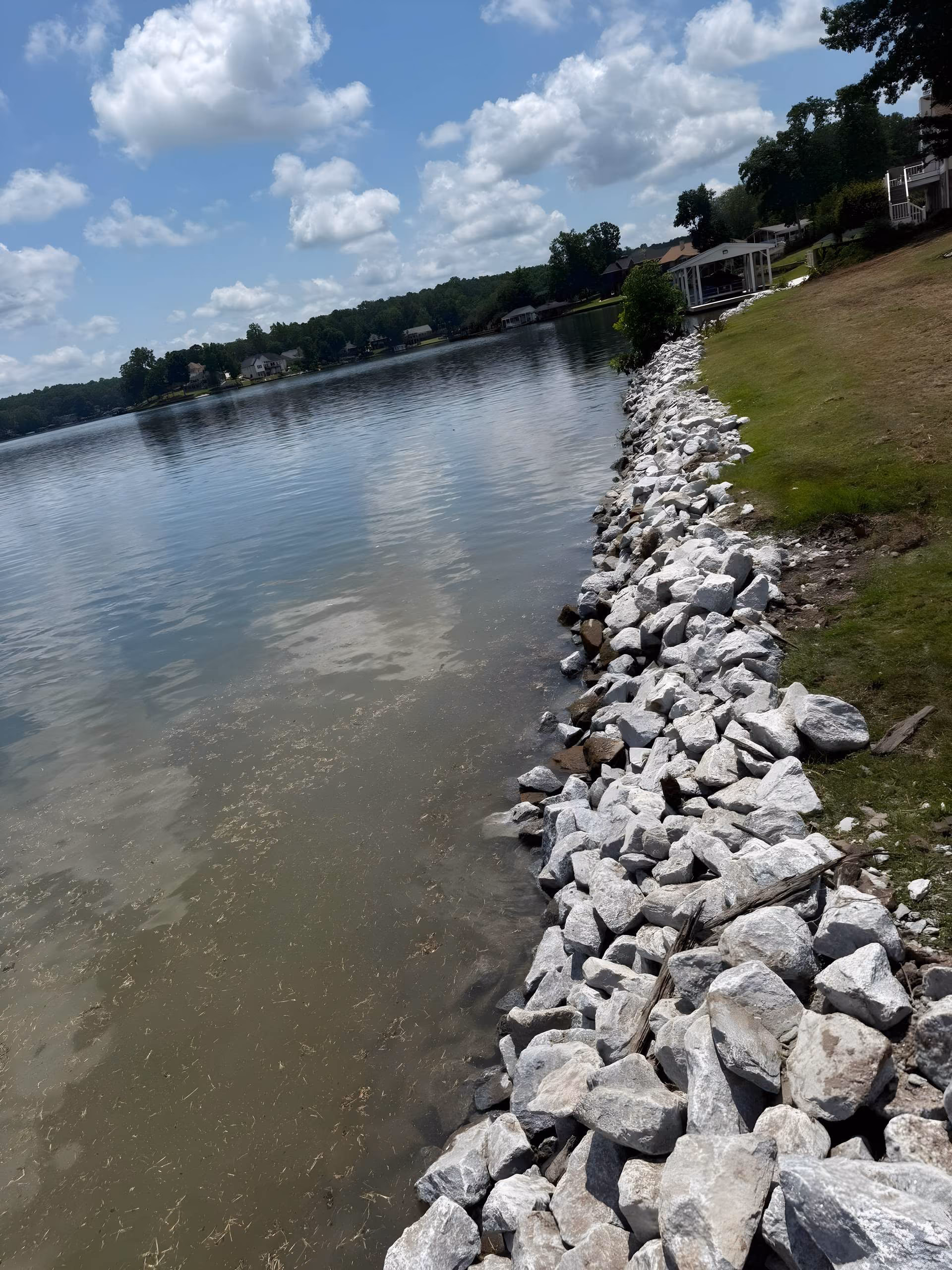 Finished riprap shoreline of stacked gray stone running along a lake bank, houses and docks in the distance