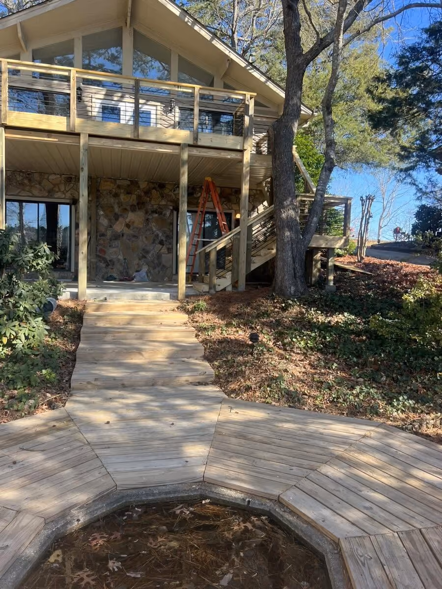 Cable-rail deck above and a new wood boardwalk below at a chalet-style home, the boardwalk wrapping an empty garden pond basin